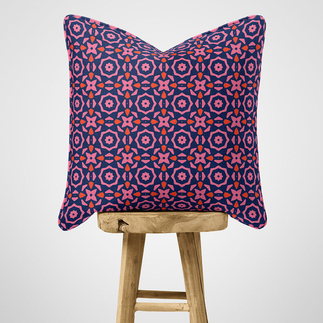 Cotton cushion with a pink red and blue geometric pattern on a wooden stool against a light grey background